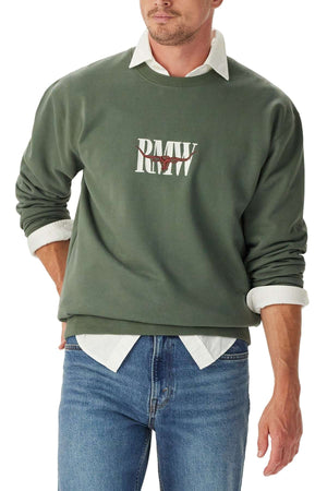A man wears the RM Williams Heritage Crew Sweatshirt in green with "RM W" in white on the front, styled over a white collared shirt and blue jeans. He has his sleeves rolled up and stands with one hand in his pocket.
