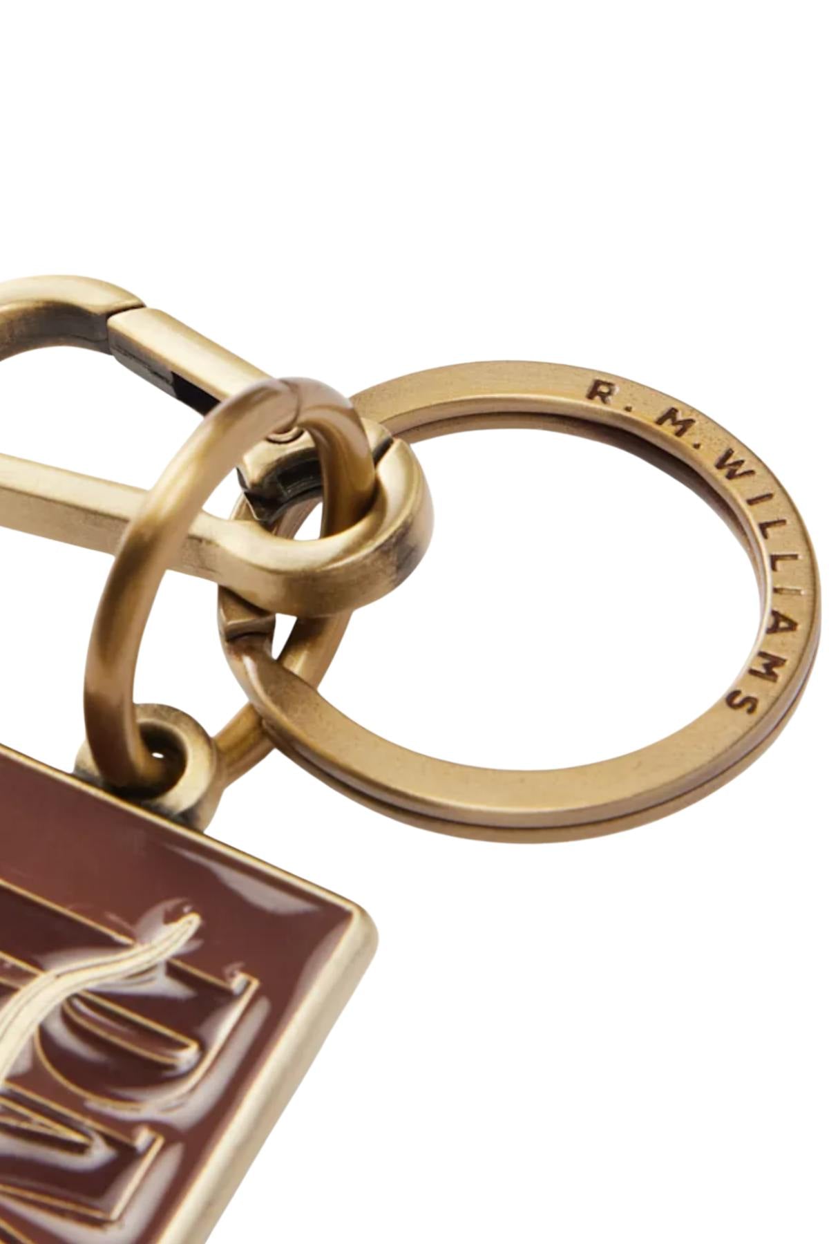 Close-up of the RM Williams Heritage Logo Enamel Key Ring, engraved with "R. M. WILLIAMS," attached to a carabiner and a brown rectangular tag featuring gold accents and sturdy brass hardware.