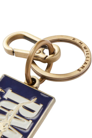 A close-up of the RM Williams Heritage Logo Enamel Key Ring, featuring “R. M. Williams” engraved on the brass ring, attached to a blue and white enamel keychain with a clasp for securing to keys or bags.