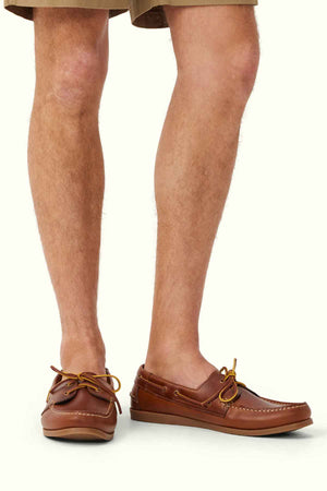 A person's lower legs and feet are shown wearing RM Williams Islet Moccasins, crafted from light brown leather and featuring tan laces. The individual is also dressed in tan shorts. The background is plain and light-colored.