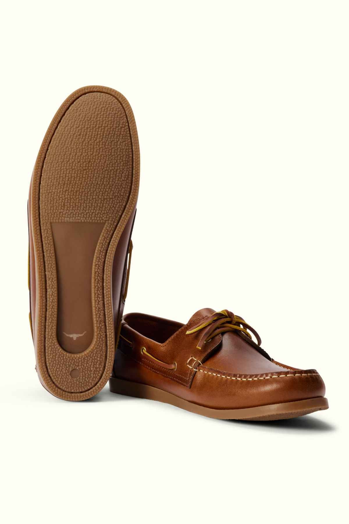 A pair of RM Williams Islet Moccasin brown leather boat shoes is displayed. One shoe stands upright, showcasing its side and top, while the other is positioned to show the textured sole. Both shoes feature leather laces and intricate stitching details.