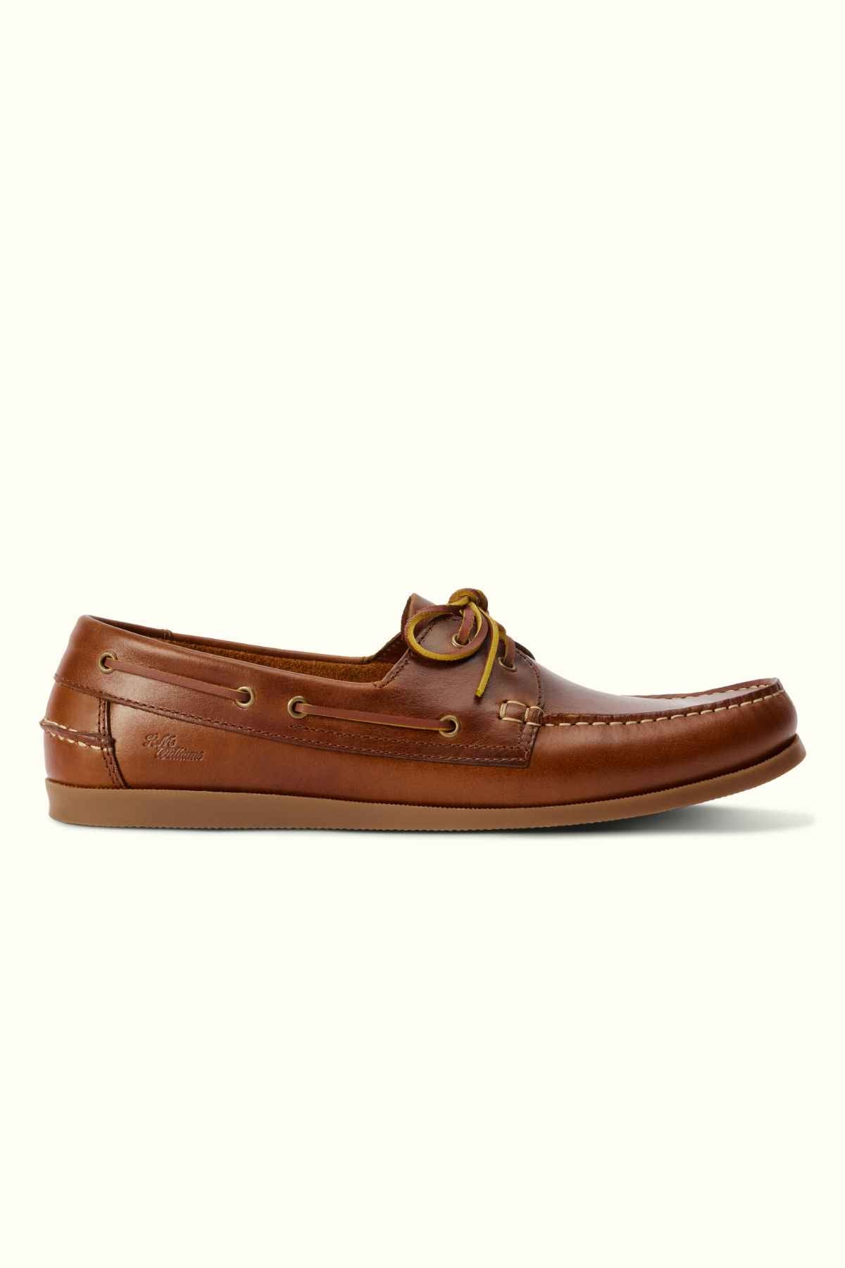 A single RM Williams Islet Moccasin in brown leather is displayed against a plain light background. The shoe features lace detailing along the sides and has a low heel with a slightly elongated toe.