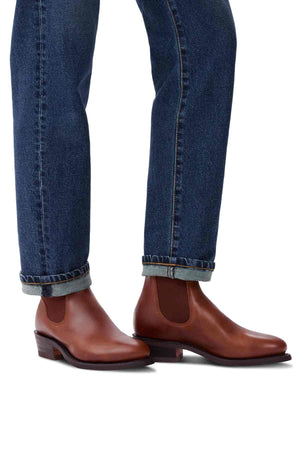 A person wearing RM Williams Lady Yearling Rubber Sole Boots with dark denim jeans slightly rolled at the cuffs. These Spanish pull-up leather Chelsea boots have a low heel and dark side panels, set against a plain white surface.