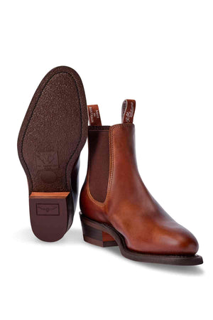 The RM Williams Lady Yearling Rubber Sole Boot features brown Spanish pull-up leather with dark elastic side panels and a back pull tab. One boot is upright, while the other shows the sole with logo branding for a comfortable and stylish look.