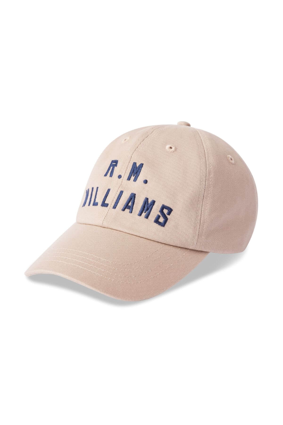 The RM Williams Logo Cap is a beige cotton twill cap with "R.M. WILLIAMS" embroidered in navy blue on the front. It features a curved brim, ventilation eyelets, and an adjustable antique brass clasp.