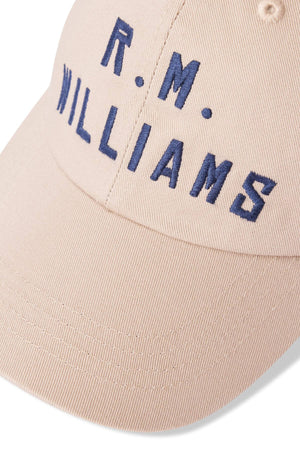 Close-up of the RM Williams Logo Cap, a cotton twill cap in beige, with "R.M. WILLIAMS" elegantly embroidered in dark blue on the front and an antique brass clasp for closure.