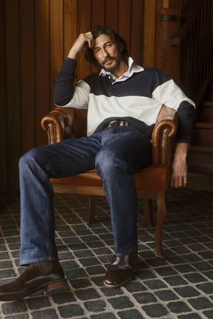 A man with wavy hair and a beard sits in a relaxed pose on a leather armchair, resting his elbow on the armrest and his hand on his head. He is wearing an RM Williams Magnus Rugby jersey made of organic cotton, paired with blue jeans and brown shoes. Behind him, there is a wooden wall and staircase.