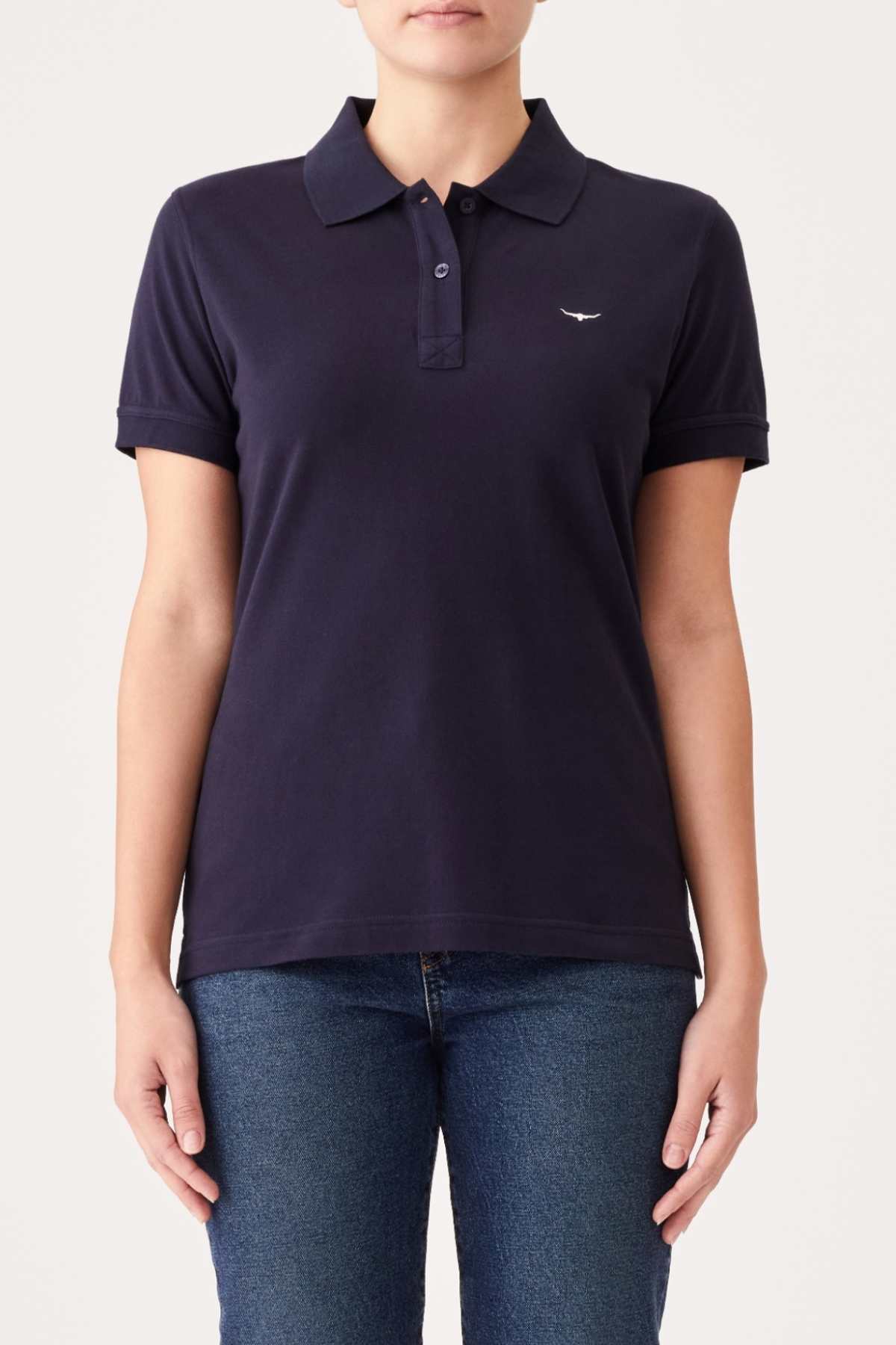 A person is wearing the RM Williams Margaret Polo, a short-sleeved, navy blue cotton polo shirt with a small logo on the left chest. They are also dressed in blue jeans. The background is plain white, and the person's face is not visible.