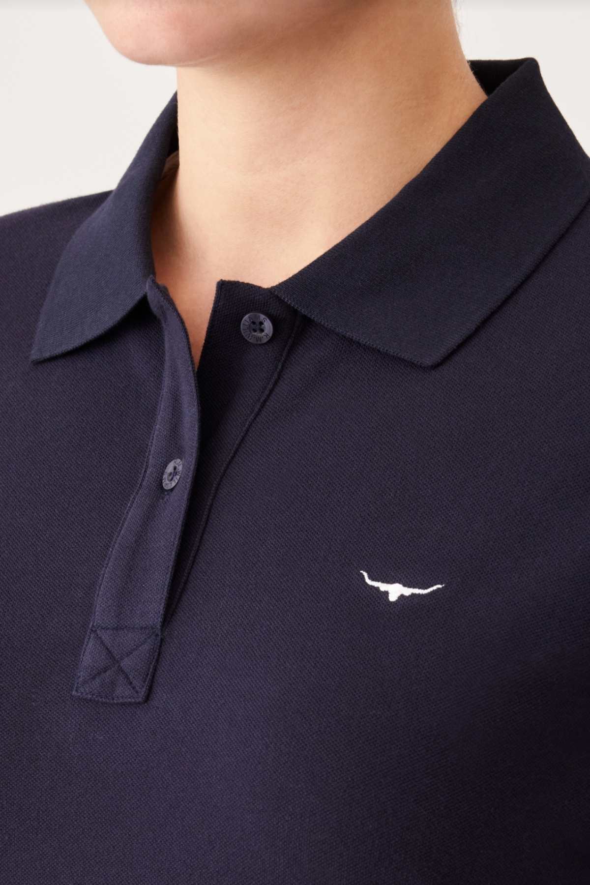 Close-up of a person wearing an RM Williams Margaret Polo. The dark-colored shirt, made from soft organic cotton, features a collar and short sleeves. It has two buttons and displays a small embroidered longhorn bull logo in white on the left side of the chest. The background is neutral.