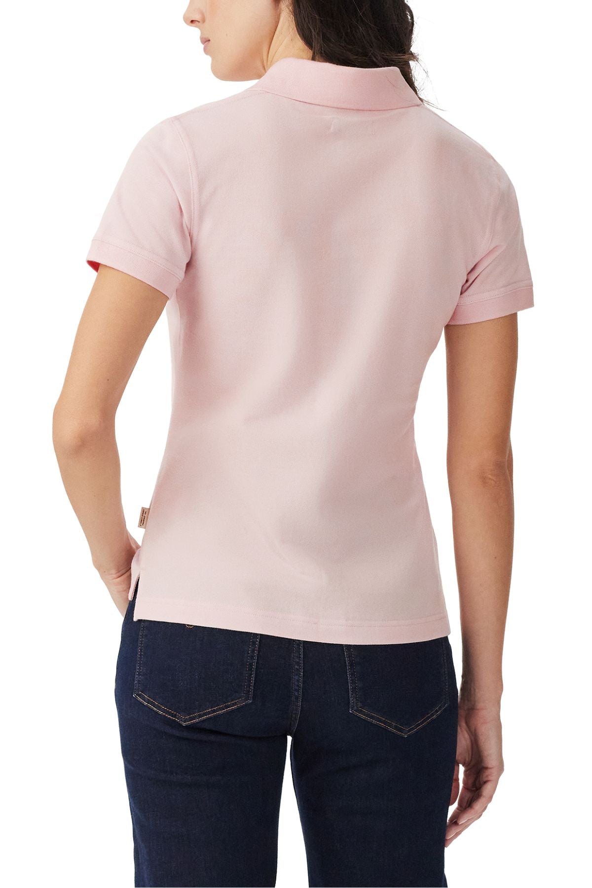 A woman is seen from behind wearing the RM Williams Margaret Polo in light pink with dark blue jeans, one hand partly in her pocket, standing against a white background.