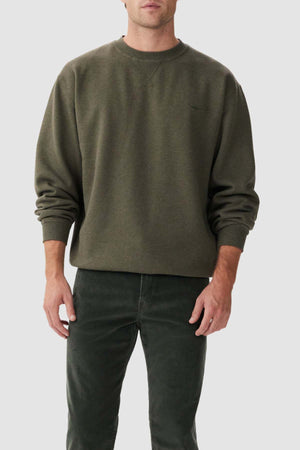 A person wearing an RM Williams Mini Longhorn Sweatshirt, which is a loose-fitting, dark green crew neck fleece sweatshirt with long sleeves, stands against a plain, light gray background. They are also dressed in dark green pants. The person's hands are relaxed by their sides and only the upper part of their body is visible.