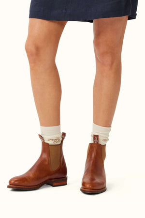 A person stands wearing the RM Williams Moriarty Rubber Sole Boot in brown leather with almond toes, paired with cream-colored socks featuring a subtle pattern. The lower part of a dark skirt is visible above the knees against a solid light background.