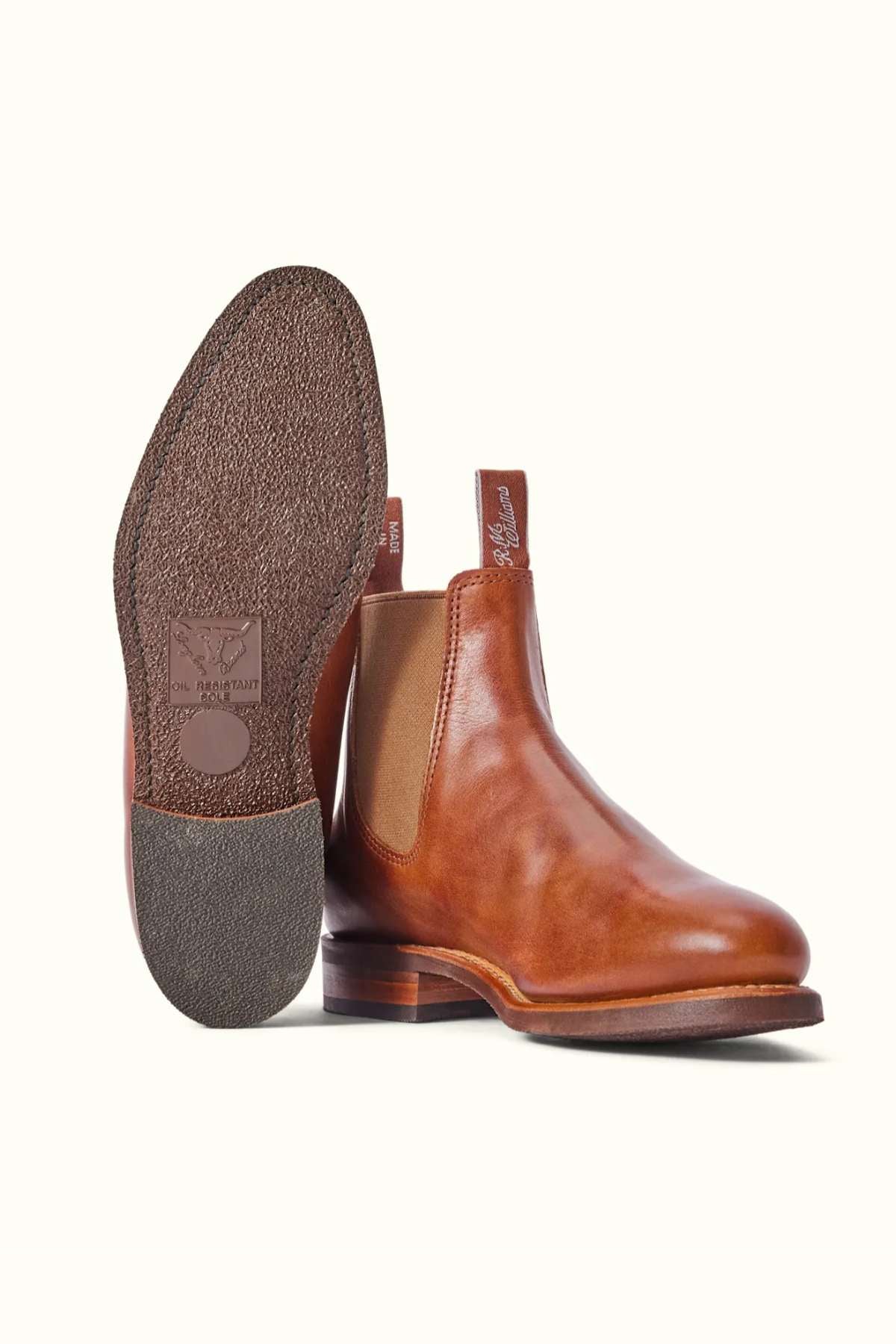 Introducing the RM Williams Moriarty Rubber Sole Boot: These tan pull-up leather Chelsea boots feature elastic side panels and a stylish design. One boot stands upright, highlighting its polished finish and almond-shaped toe, while the other displays the textured sole adorned with the brand logo. Both boots are equipped with pull tabs featuring embossed lettering, ensuring ease of wear without compromising on style.