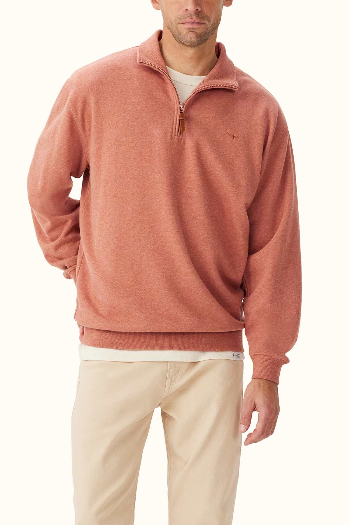 A man wearing the RM Williams Mulyungarie Fleece in a rust color stands against a plain light background. This quarter-zip sweater, crafted from fine French organic cotton terry, is layered over a white shirt and beige pants and features a subtle logo on the chest.