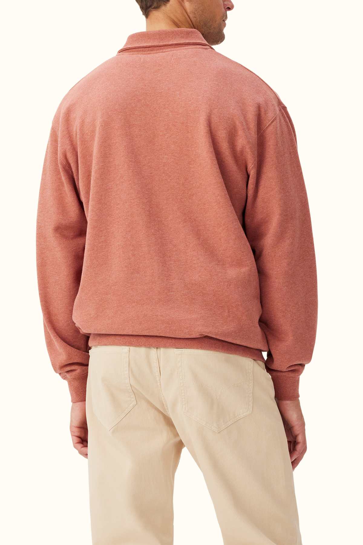 A person wearing the RM Williams Mulyungarie Fleece in rust-colored organic cotton stands with their back to the camera, paired with light beige pants. The plain off-white background accentuates the understated elegance of the French cotton terry material.