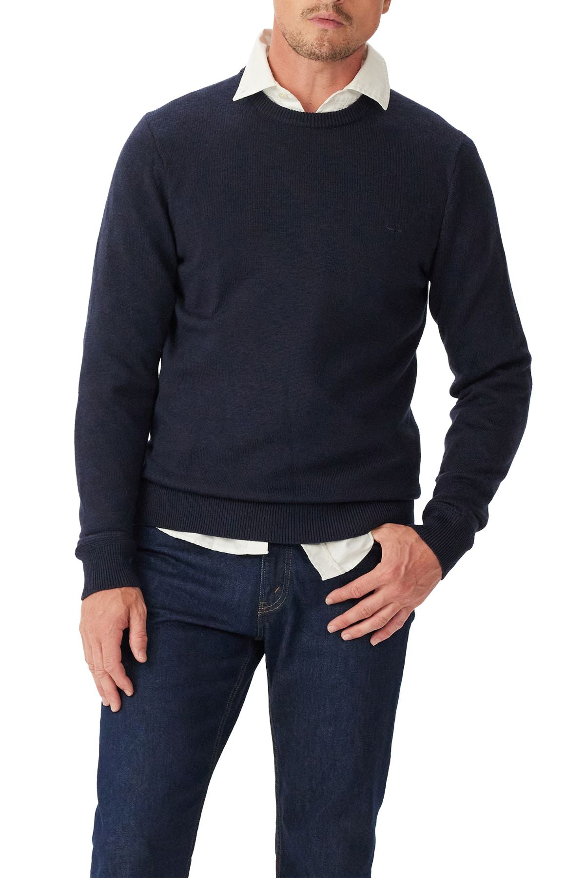 A man wearing the RM Williams Murphy Merino Crew Knit in navy over a white collared shirt and dark blue jeans stands with one hand in his pocket against a white background.