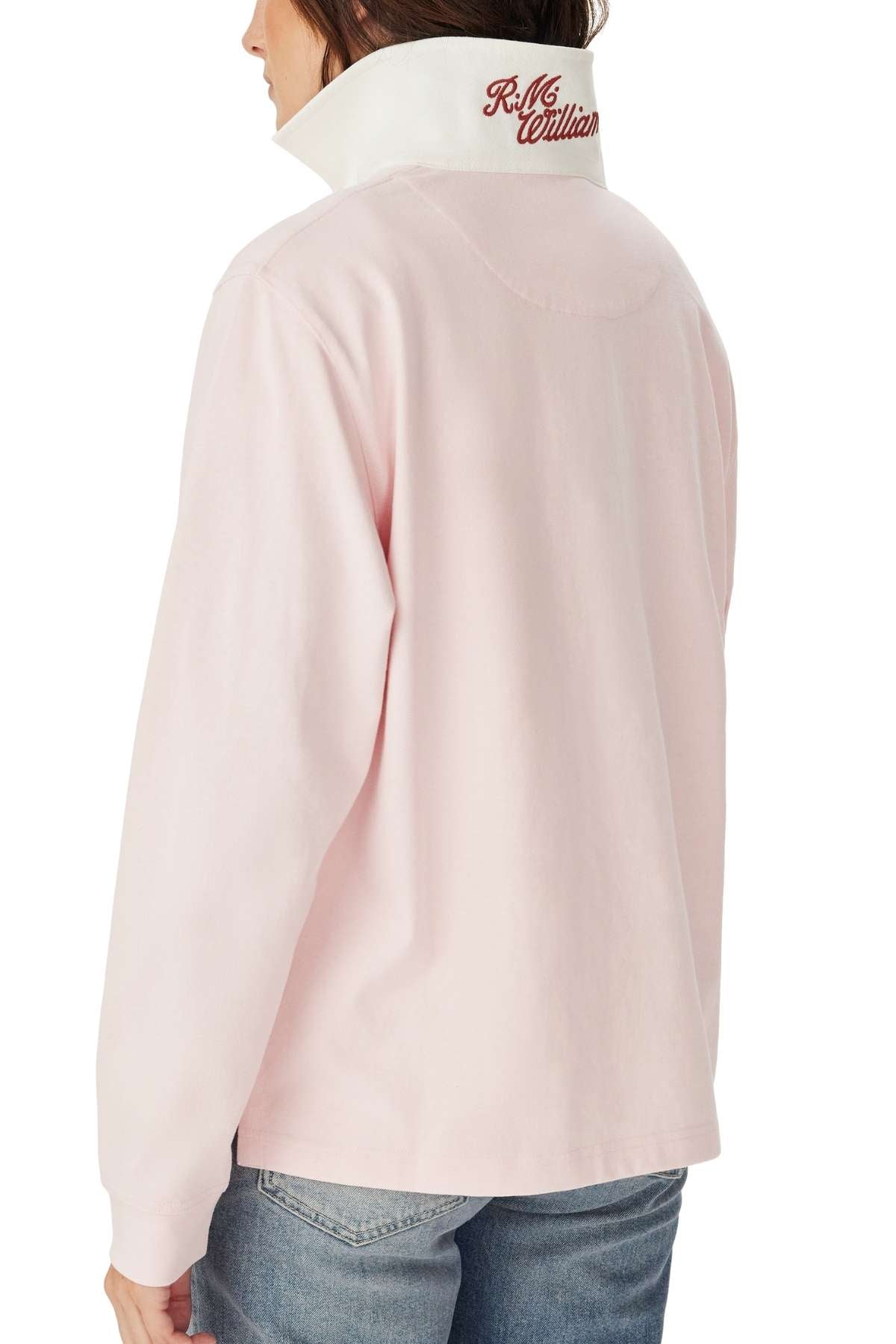 The person is wearing the RM Williams Narran Heritage Rugby, a light pink long-sleeve shirt with a white collar and red embroidered text on the collar, seen from the back and paired with blue jeans.