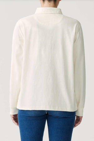 The image features an individual dressed in the RM Williams Narran Rugby, a long-sleeved, off-white shirt crafted from organic cotton, paired with blue jeans. Standing with their back to the camera against a plain background, they exude casual elegance.