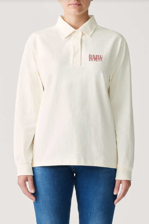 A person wearing the RM Williams Narran Rugby, a cream long-sleeve jersey made from organic cotton, showcases "DREW" embroidered in red on the chest and is styled with blue jeans. This shirt includes a classic collar and buttons, set against a plain, light-colored background.