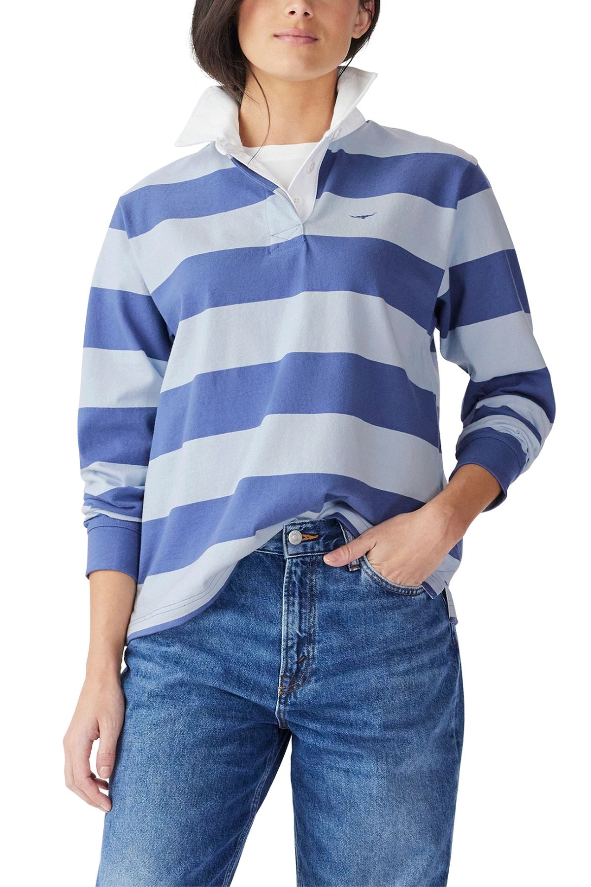A person wearing the RM Williams Narran Stripe Rugby, a blue and light blue striped organic cotton shirt with a brushed twill collar, and blue jeans stands with one hand in their pocket. Their face is partially visible.