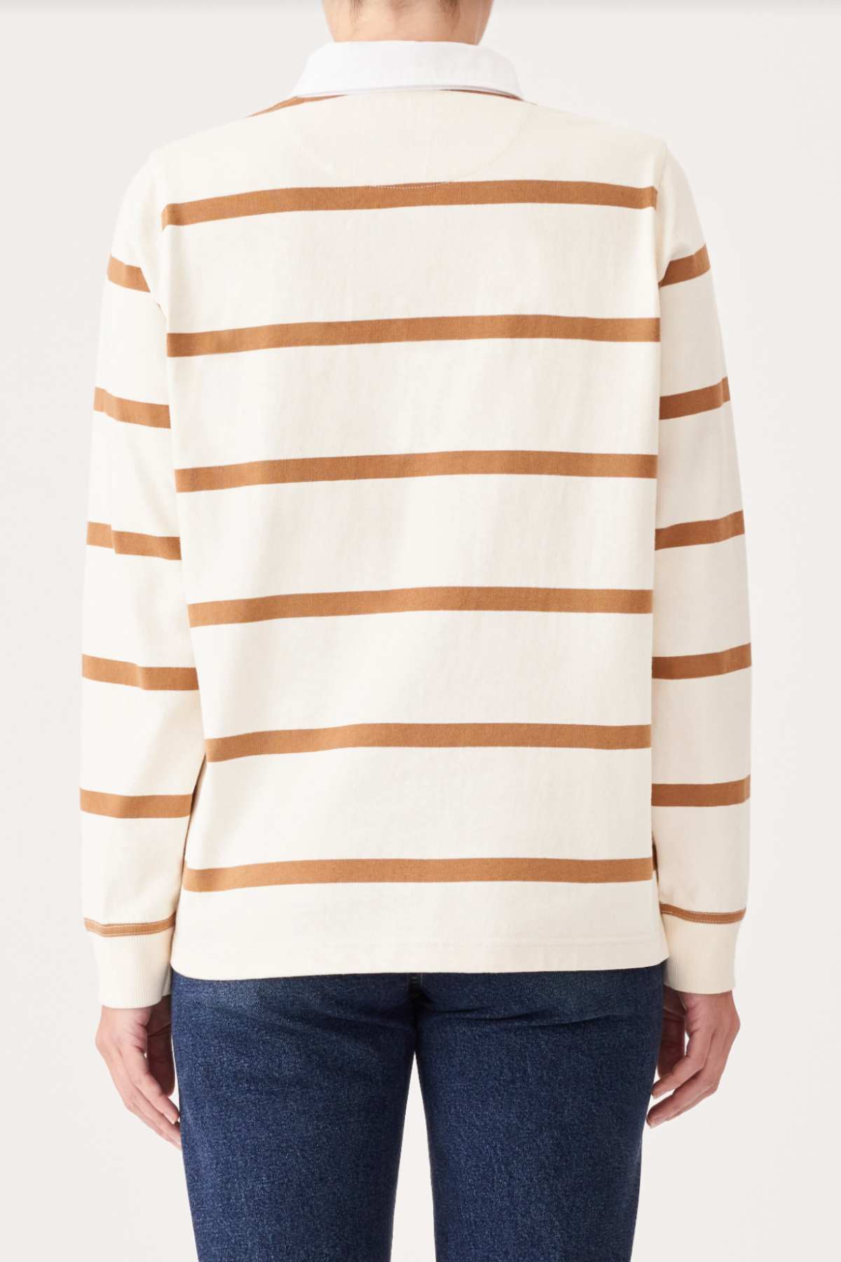 A person wearing the RM Williams Nundle Rugby, a long-sleeve, cream-colored sweater featuring horizontal brown stripes, is facing away with their back to the camera. They are also wearing dark blue jeans against a plain and light-colored background.