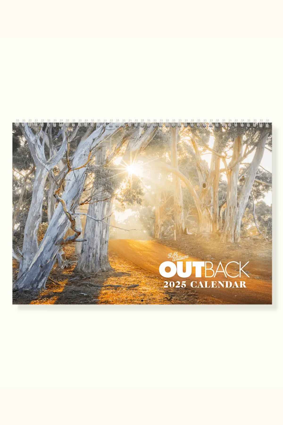The RM Williams Outback Calendar 2025, a stunning wall calendar by gifted Australian photographers, features sunlight filtering through tall trees along a dirt path. The warm glow casts long shadows, creating a serene and picturesque outback scene.