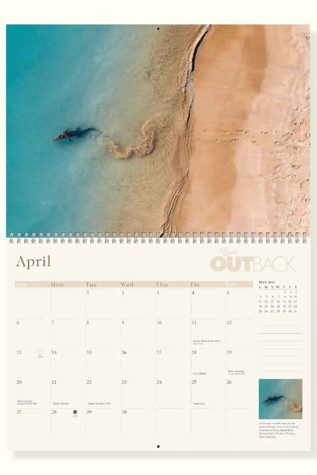 The top half of the RM Williams Outback Calendar 2025 showcases an aerial view of sandy beach and turquoise waters by Australian photographers, with dates and marked events below, bound by a spiral at the top.