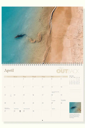 The top half of the RM Williams Outback Calendar 2025 showcases an aerial view of sandy beach and turquoise waters by Australian photographers, with dates and marked events below, bound by a spiral at the top.