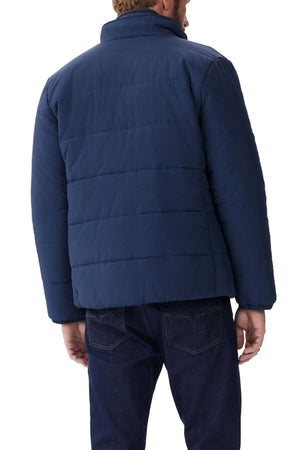 A person is wearing a RM Williams Padstow Jacket and dark jeans. The rear view highlights the blue jacket's padded texture, made from recycled nylon with Primaloft fill, and its simple design against a plain white background.