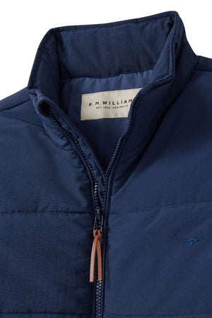 Close-up of the RM Williams Padstow Jacket, a dark blue puffer with Primaloft fill and high collar. Features a brown leather zipper pull and an embroidered longhorn logo on the chest. Made from recycled nylon with an inside label: "R.M. Williams, Est. 1932, Australia.