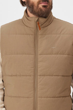 A bearded man wears the RM Williams Padstow Vest, a tan puffer made from recycled nylon, over a light beige shirt; it offers lightweight comfort, a high collar, front zipper, and features a small embroidered chest logo.
