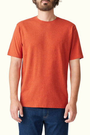 A person wearing an RM Williams Parson T-Shirt in orange organic cotton and blue jeans stands against a plain light background. The T-shirt has short sleeves and a crew neck, while the person's hands are relaxed by their sides.