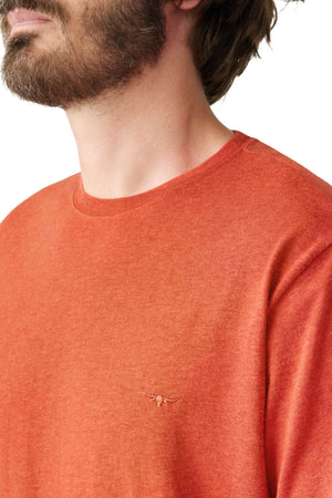 A person with a beard is wearing the RM Williams Parson T-Shirt, an orange, organic cotton crew neck top featuring a small embroidered horns logo on the chest. The image is cropped to show the upper body and part of their face, highlighting its casual style.