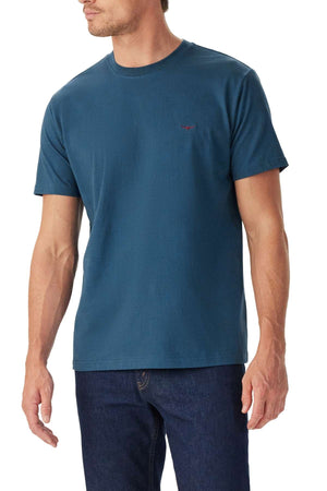 A man wearing the RM Williams Parson T-Shirt in plain teal with dark blue jeans stands slightly turned against a white background.