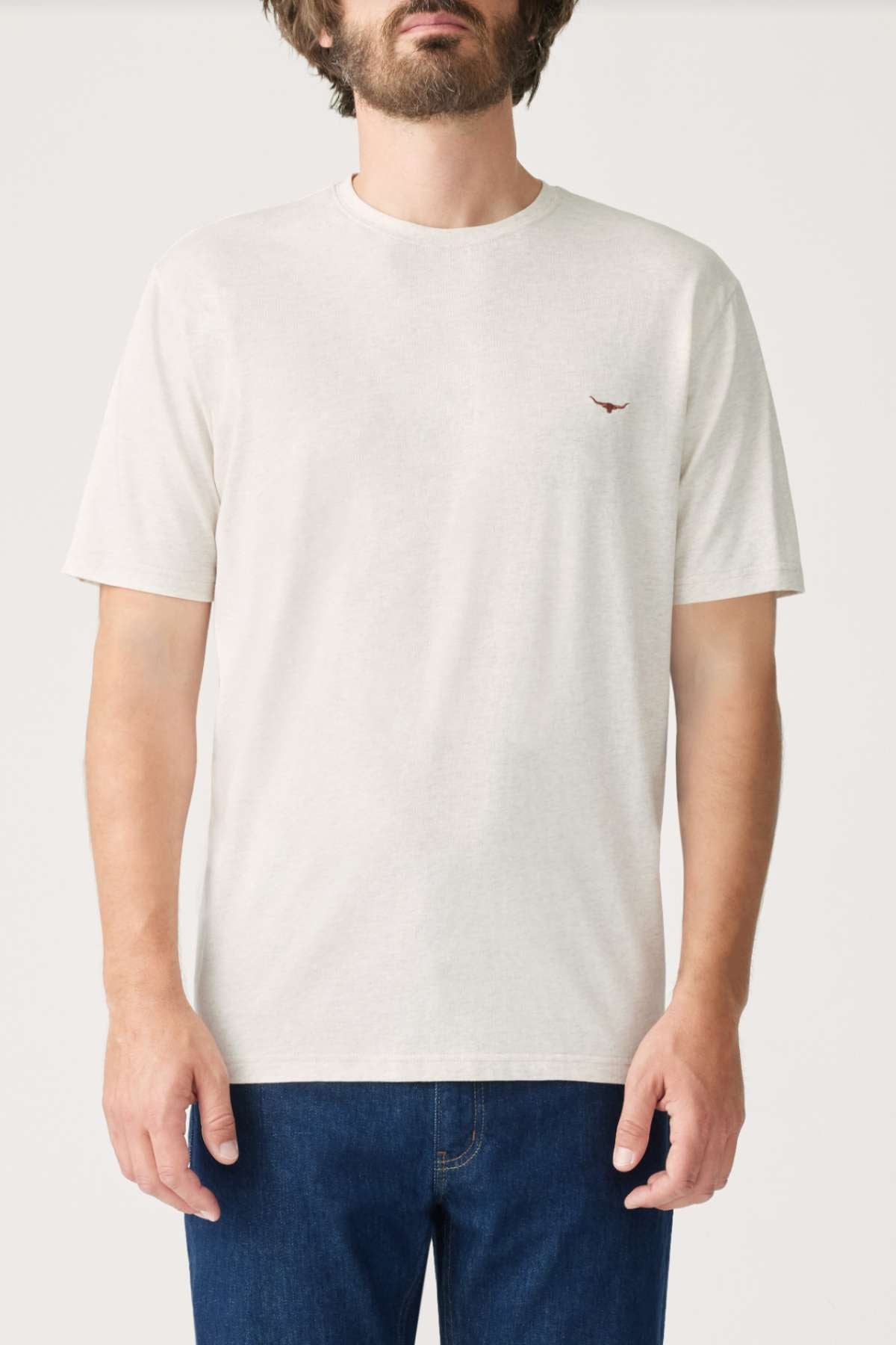 A person is wearing a simple and casual RM Williams Parson T-Shirt, made of mid-weight cotton jersey with a small Longhorn embroidery on the chest, paired with blue jeans against a neutral background. The plain white T-shirt is perfect for everyday wear.
