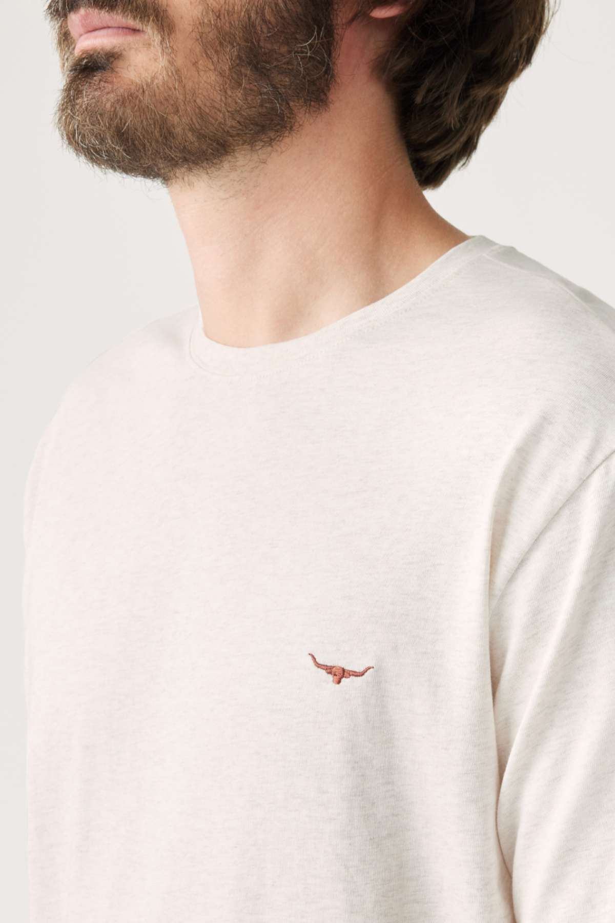 A person is seen wearing a cream-colored RM Williams Parson T-Shirt, made from mid-weight cotton jersey and adorned with a small Longhorn embroidery on the chest. The image emphasizes the upper part of the shirt and the logo, with only part of the person's face visible.