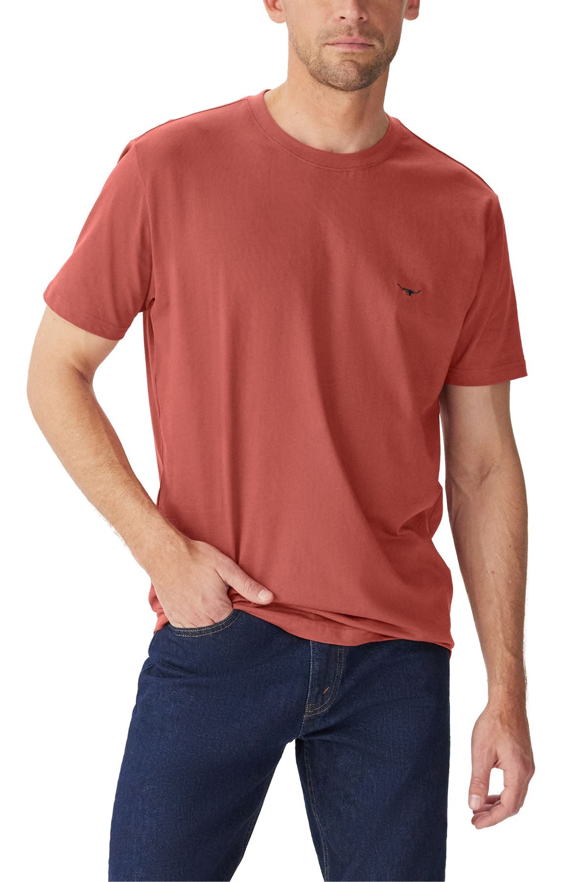 A man wearing a rust-colored RM Williams Parson T-Shirt made from organic cotton and dark blue jeans stands with one hand in his pocket against a white background.