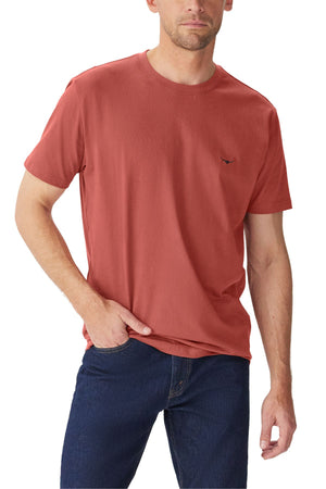 A man wearing a rust-colored RM Williams Parson T-Shirt made from organic cotton and dark blue jeans stands with one hand in his pocket against a white background.