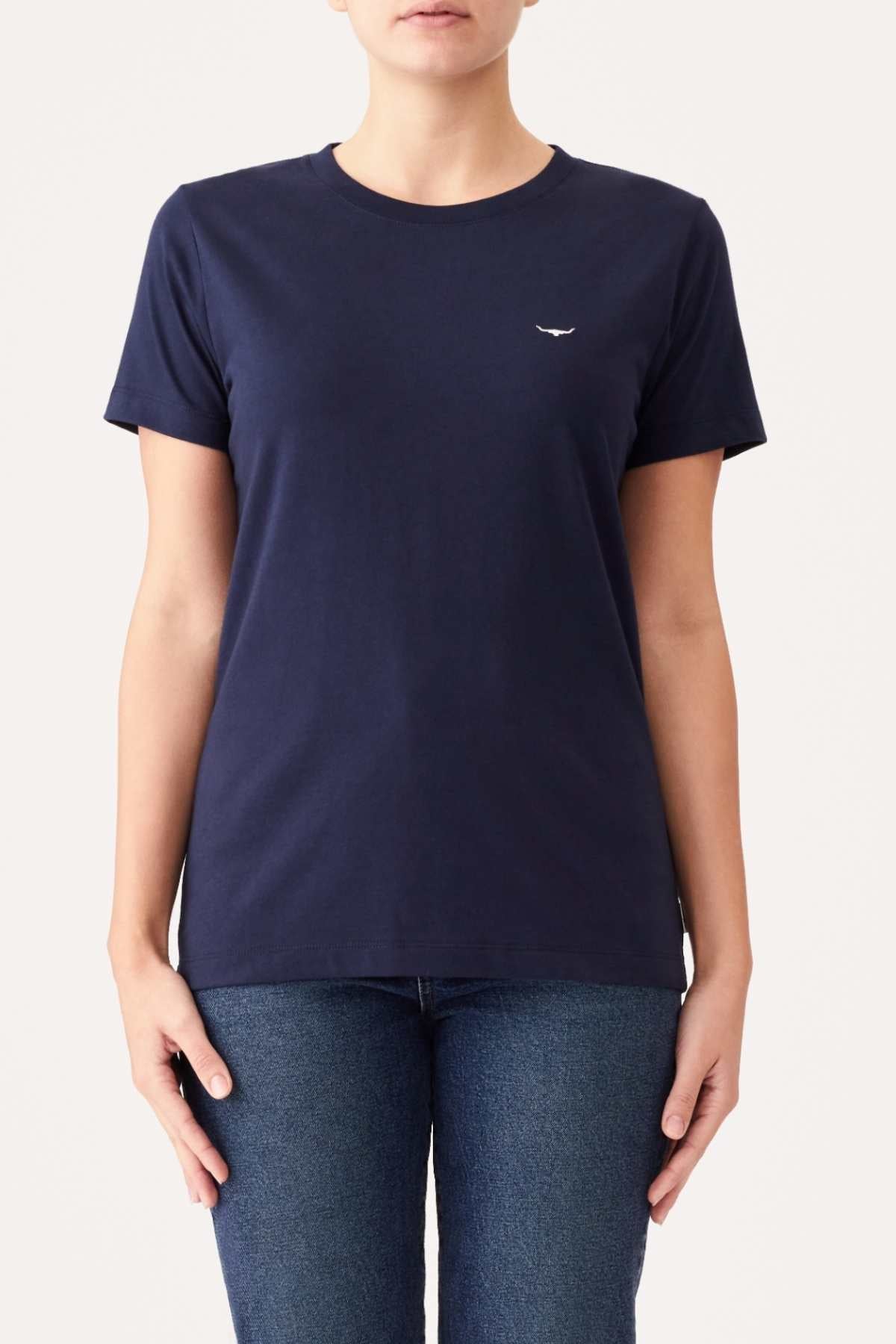 A person is wearing an RM Williams Piccadilly T-Shirt, which is plain, fitted, navy blue, and made from organic cotton with a small white logo on the left side of the chest. They are also wearing medium blue jeans. The background is a plain light color. The person’s face is not visible.