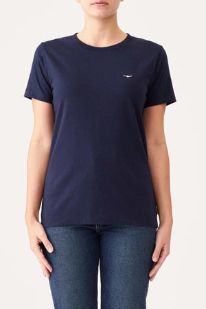 A person is wearing an RM Williams Piccadilly T-Shirt, which is plain, fitted, navy blue, and made from organic cotton with a small white logo on the left side of the chest. They are also wearing medium blue jeans. The background is a plain light color. The person’s face is not visible.