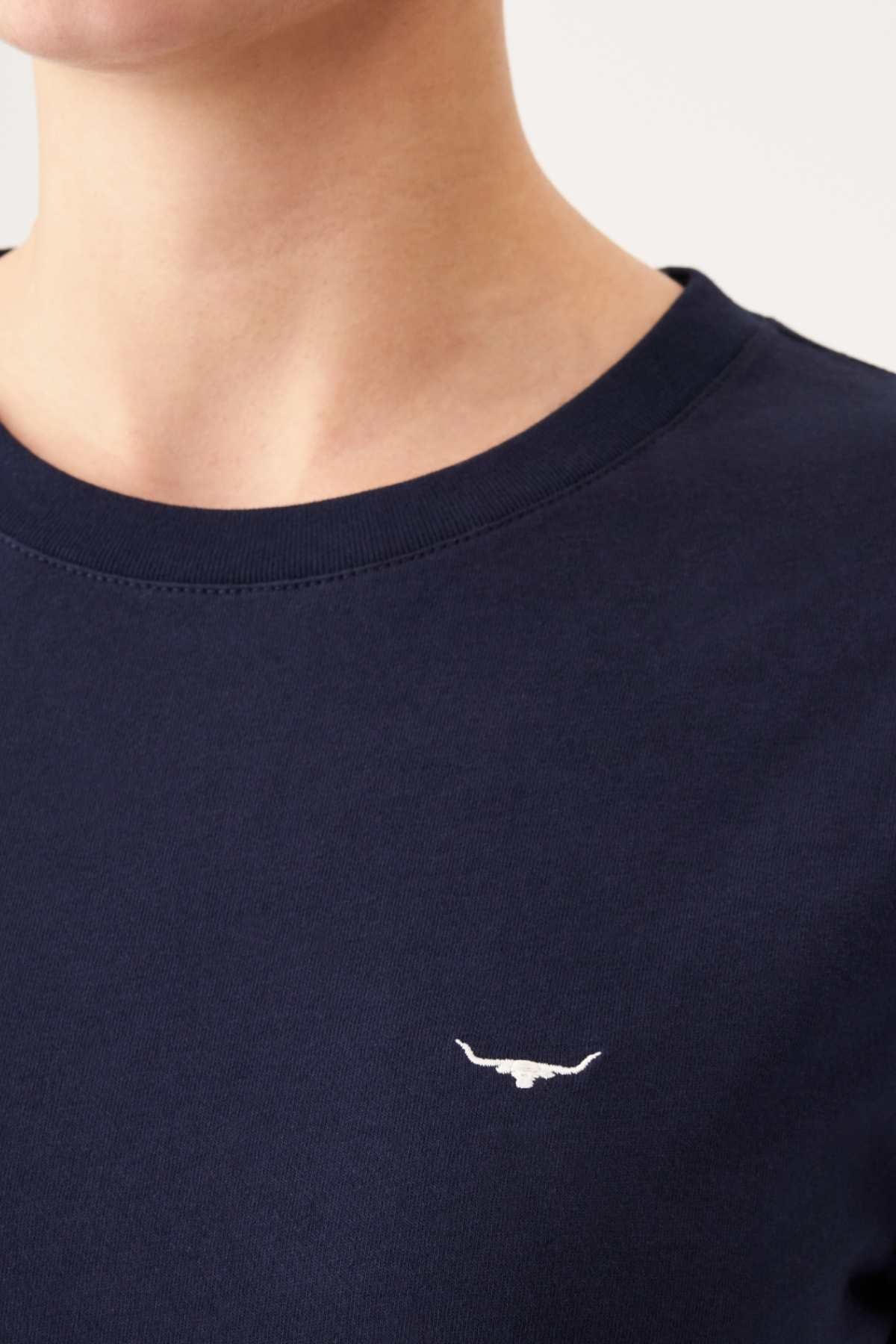 A close-up of a person wearing the RM Williams Piccadilly T-Shirt, a dark blue crew-neck shirt crafted from organic cotton. The shirt features a small embroidered white longhorn logo on the left side of the chest. The background is a plain, light-colored backdrop, with the person's face partially visible from the neck down.