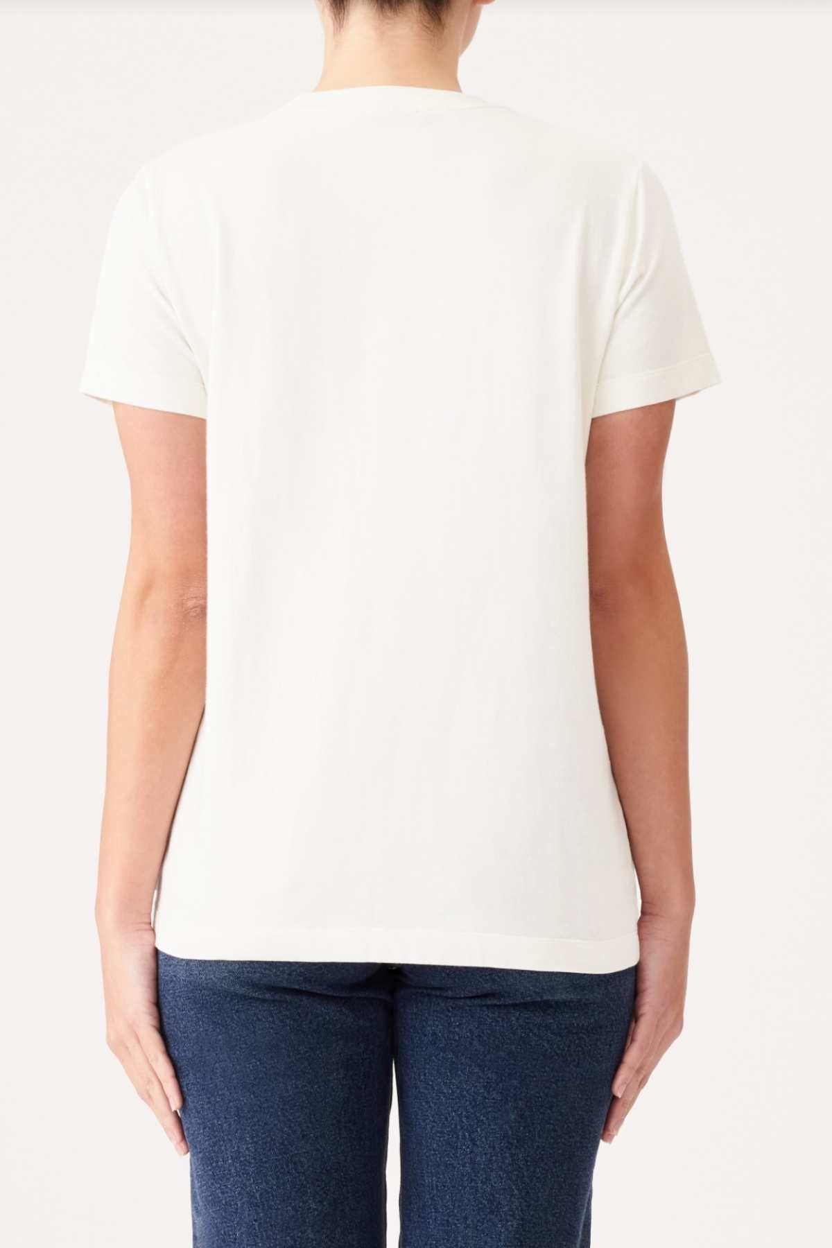 A person stands facing away from the camera, wearing a plain white RM Williams Piccadilly T-Shirt and blue jeans. The background is a simple, solid off-white color. The person's arms are relaxed by their sides.