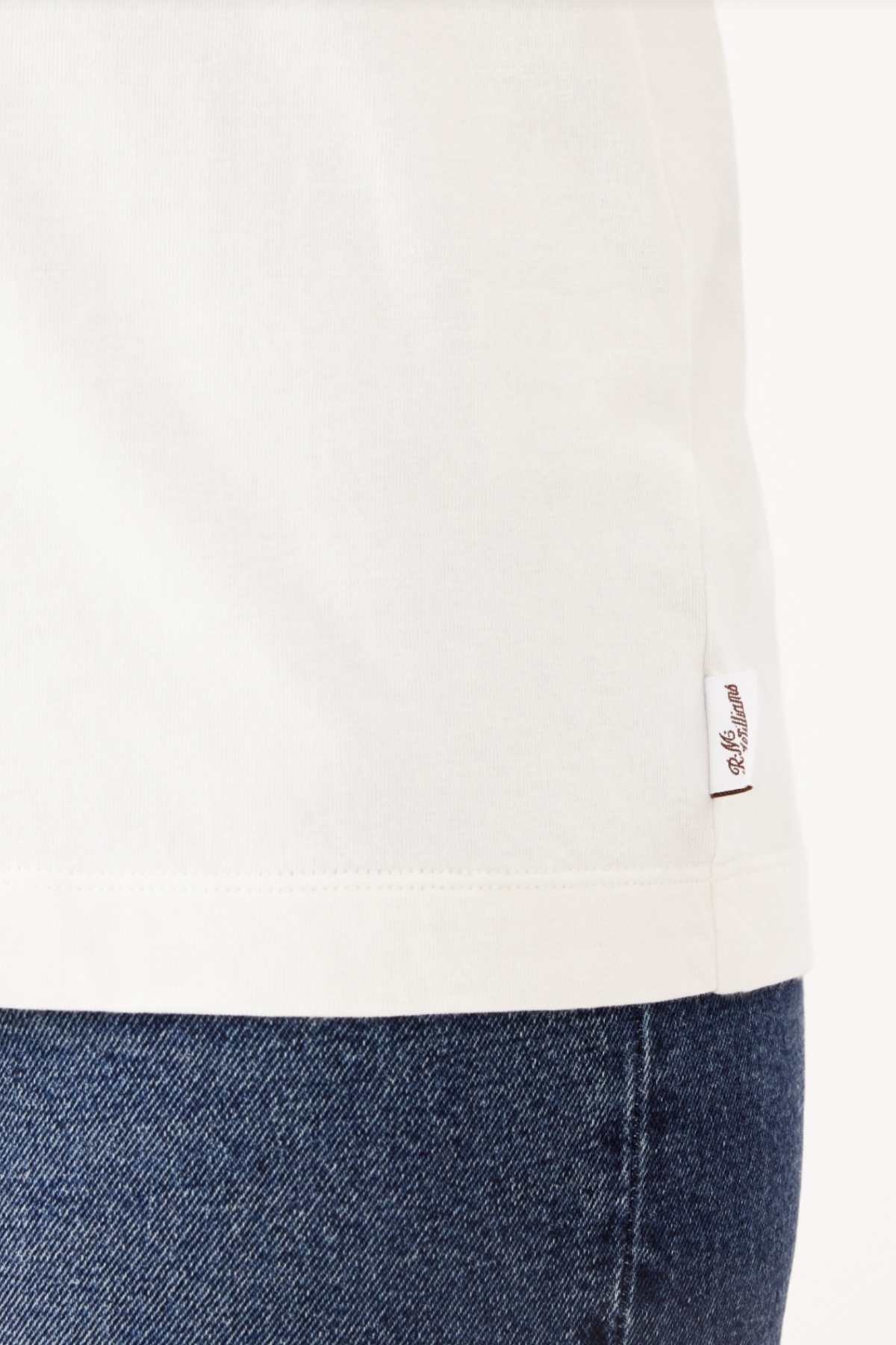 Close-up of a person wearing an RM Williams Piccadilly T-Shirt and blue jeans. The focus is on the bottom hem of this everyday T-shirt, which has a small white tag with red lettering.