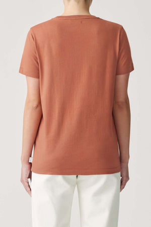 A person is seen from the back against a plain background, wearing the RM Williams Piccadilly Tee in rust with longhorn embroidery and white pants.