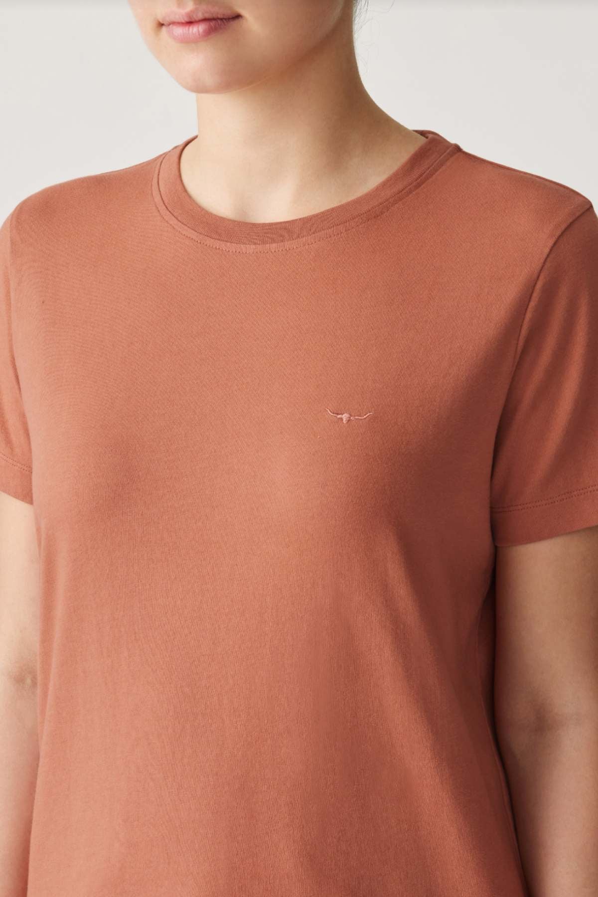 A person wearing the RM Williams Piccadilly Tee, a rust organic cotton T-shirt with a small bird logo on the chest. It features short sleeves and a round neckline against a simple, light backdrop.
