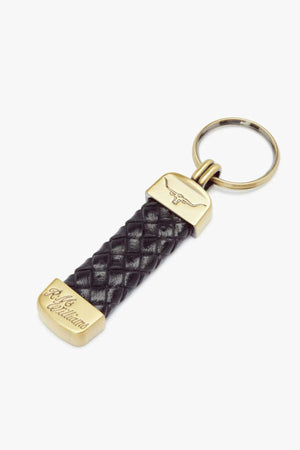 The RM Williams Plaited Key Ring includes a gold-colored metal ring and tips, a braided black kangaroo leather strap, and brass-toned fittings with engravings featuring a longhorn embossed logo and stylized text.