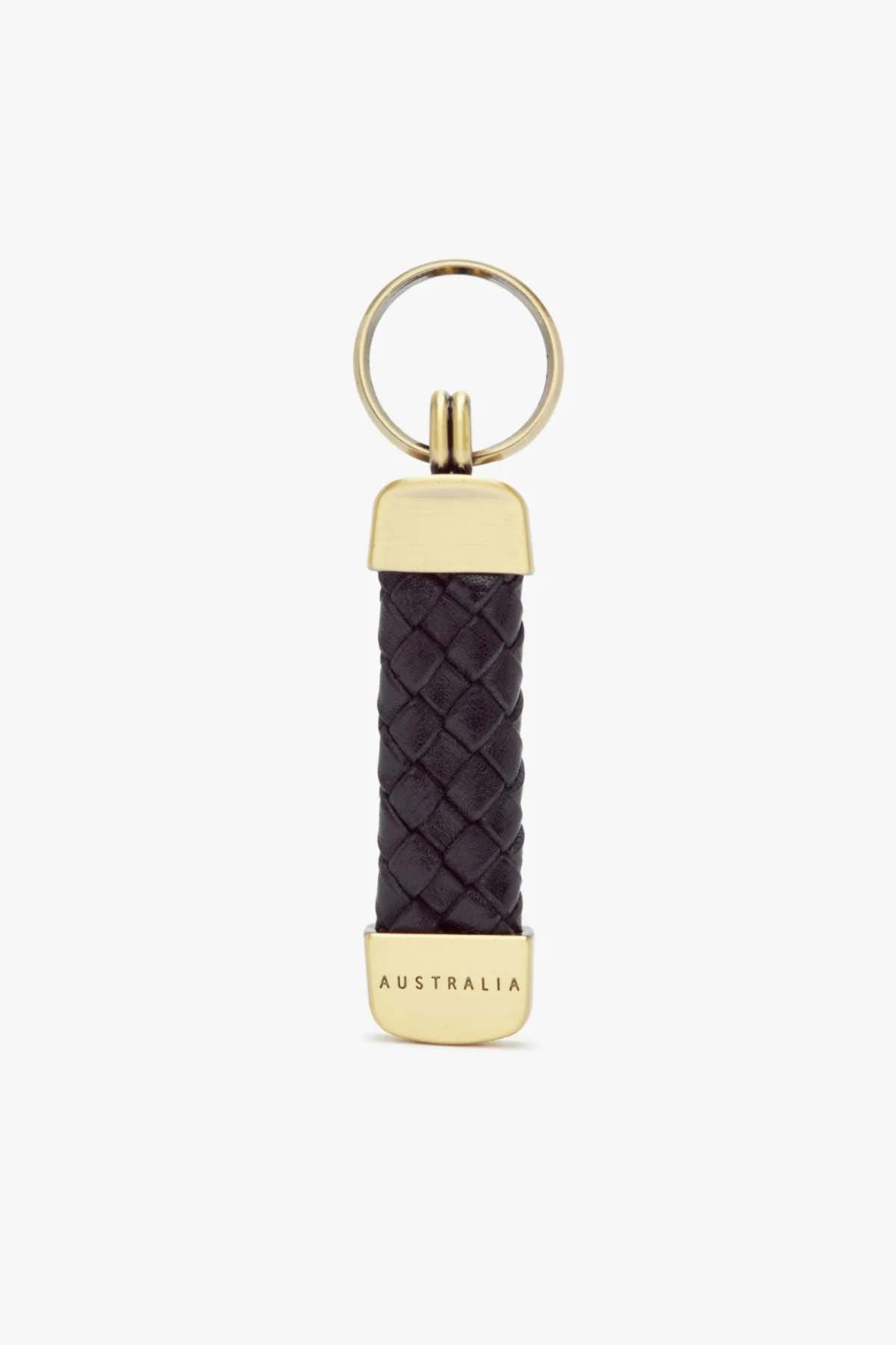 The RM Williams Plaited Key Ring features a gold ring with a woven black Kangaroo leather handle, accented by brass-toned fittings and engraved "AUSTRALIA" metal ends. The design is simple and elegant against a plain white background.