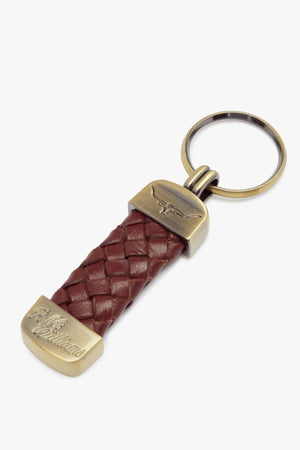The RM Williams Plaited Key Ring features a braided brown kangaroo leather strap, brass-toned fittings, and a round key ring. One metal end is engraved with text, and the other has an engraved longhorn image.