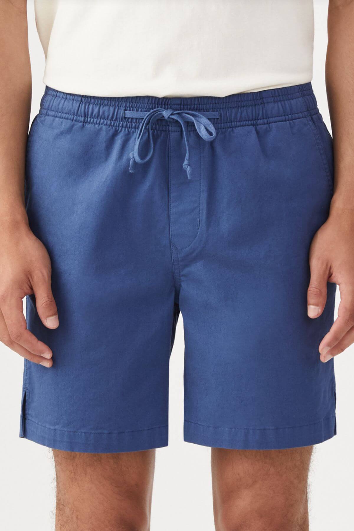 A person stands against a plain background, wearing a white shirt and RM Williams R.M.W Rugby Short in blue stretch cotton twill, shown from waist to mid-thigh with arms relaxed at their sides.
