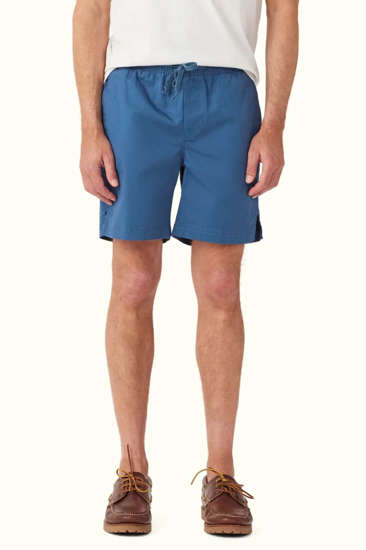 A person wearing a white shirt, RM Williams R.M.W Rugby Shorts in blue cotton with an elasticated waist and a drawstring adjuster, and brown boat shoes stands against a plain background. The shorts feature a casual, relaxed fit.